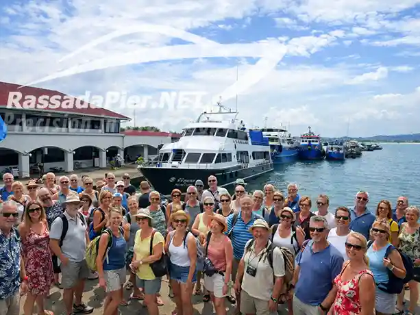 A large group of happy tourists gathering in front of a docked passenger boat at the Phuket Ferry Terminal.