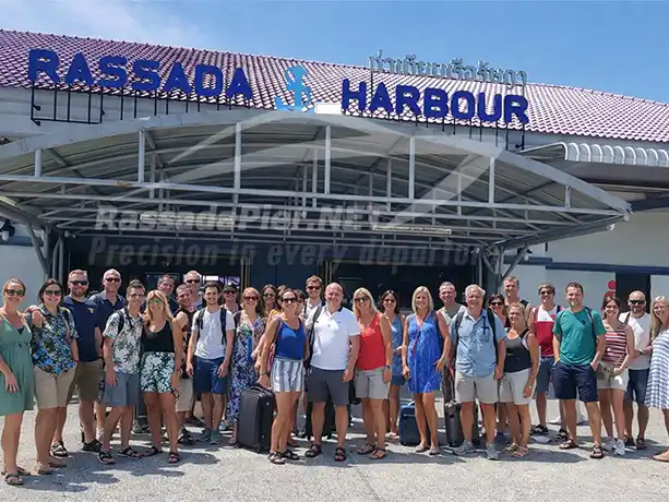 A large group of tourists with luggage posing in front of the main entrance sign at Rassada Pier Terminal in Phuket